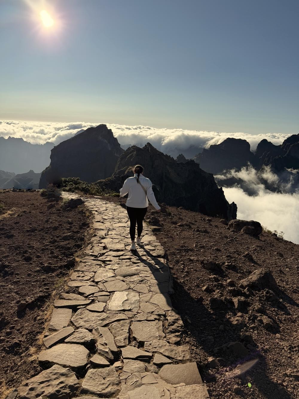 Jamie walking a stone trail on a Madeira mountain ridge above the clouds, with the sun overhead